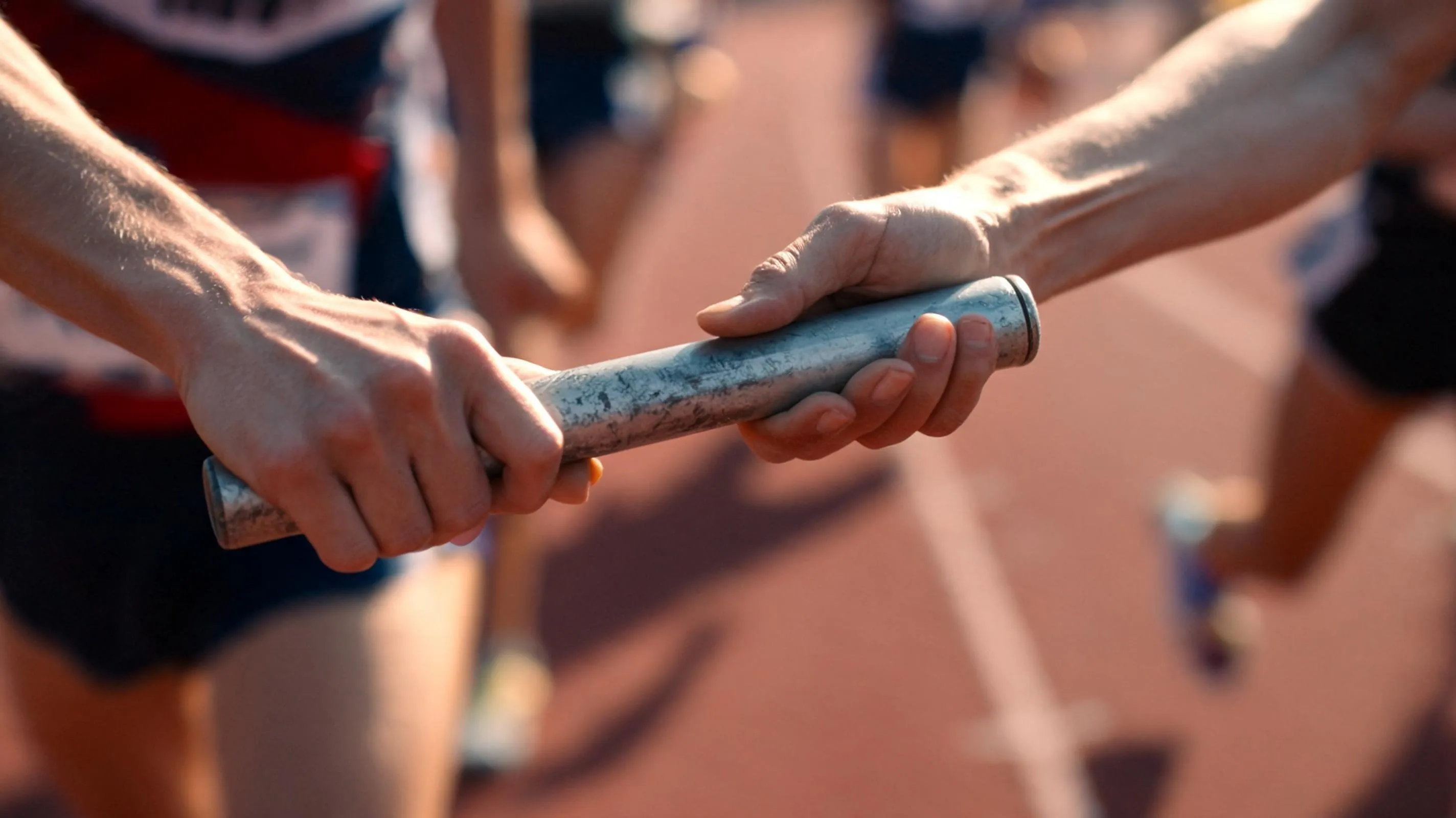 Two relay runners at the instant a baton passes from one hand to the other, lit by low golden side light
