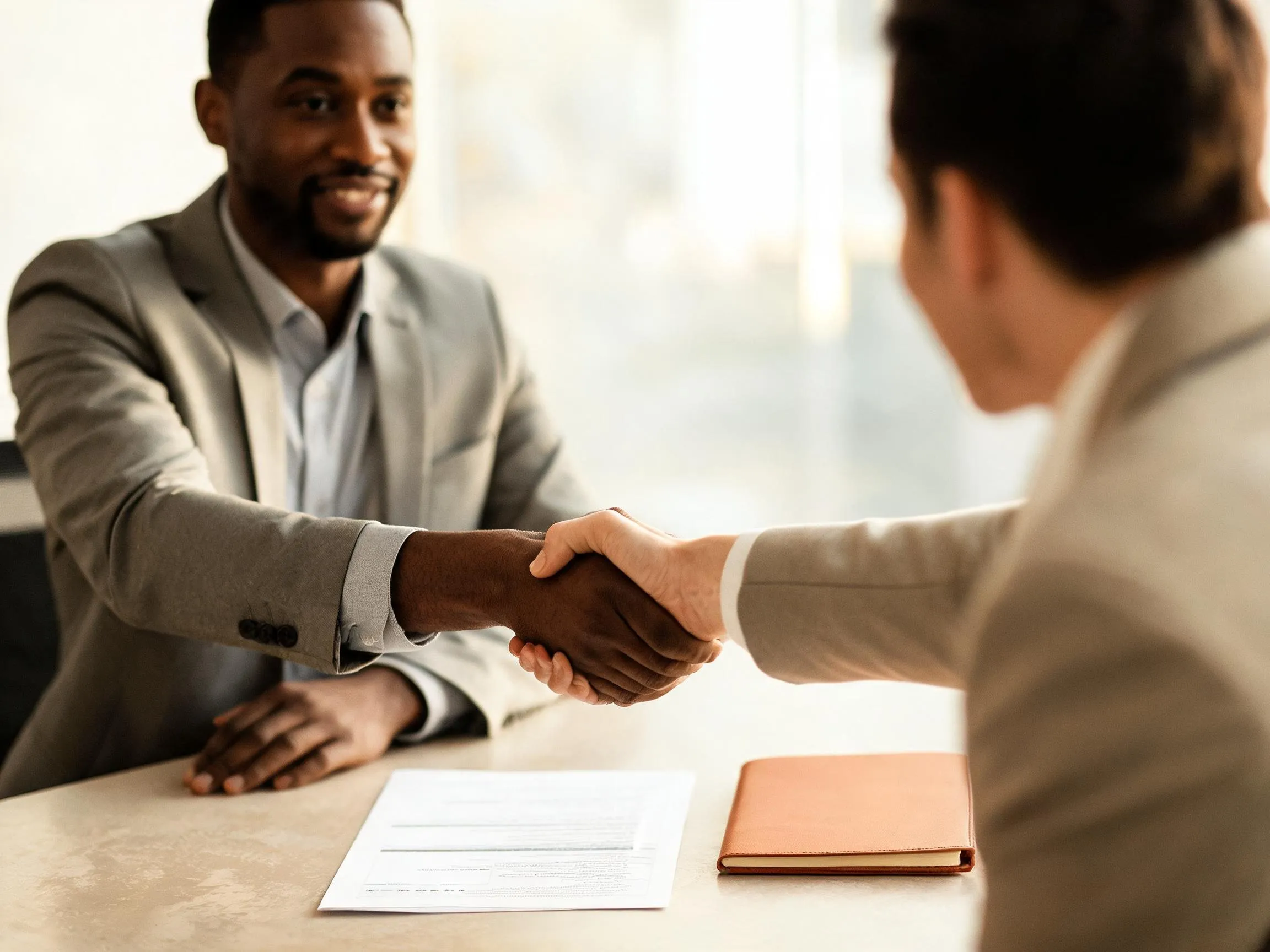 Two people shake hands across a table with signed papers and a leather folio between them